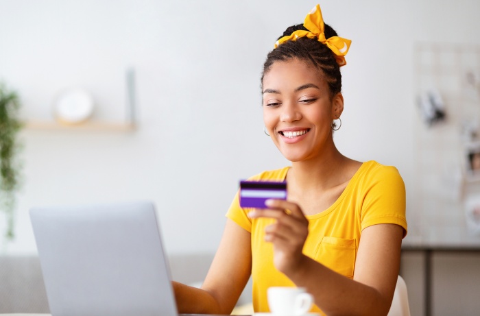 a young woman smiling, sitting at her laptop with a credit card she is using online to build her credit