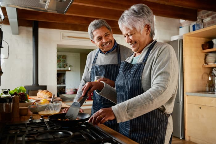 Elderly multi-ethnic couple laughing together in the kitchen. Happily retired and cooking breakfast with each other.