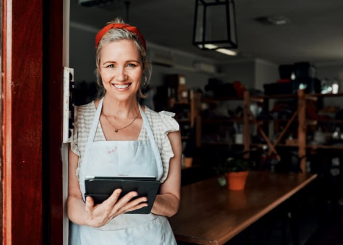 an older woman standing at the door to her business while holding an iPad