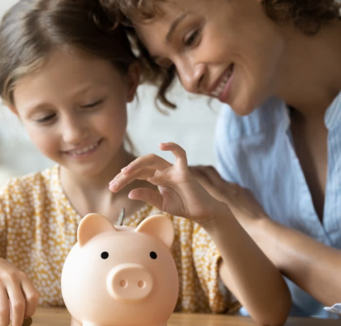 A mother teaching her daughter how to save money by placing a coin in a piggy bank